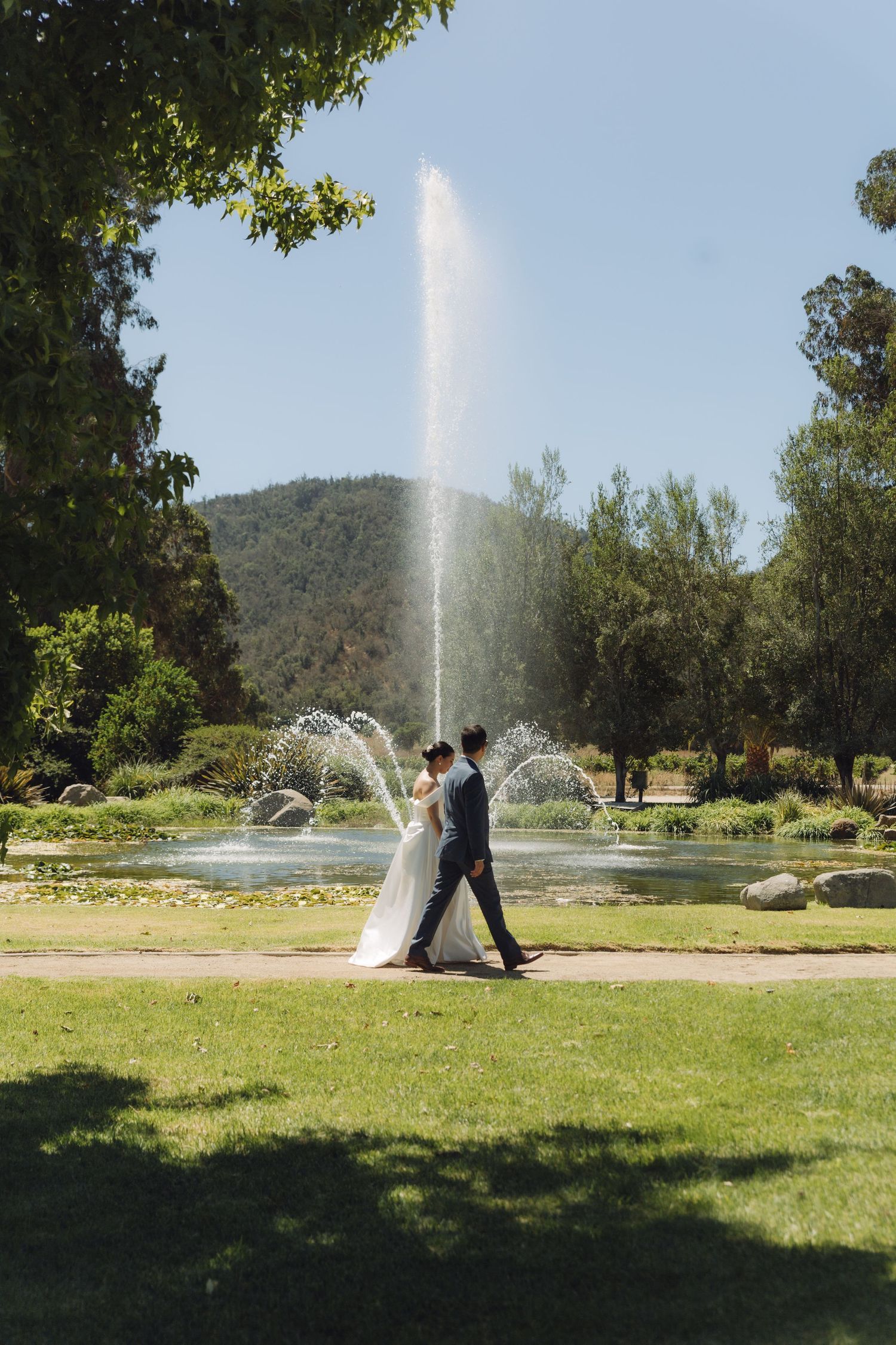 Newlyweds walking past a grand fountain in a lush green park on their wedding day.
