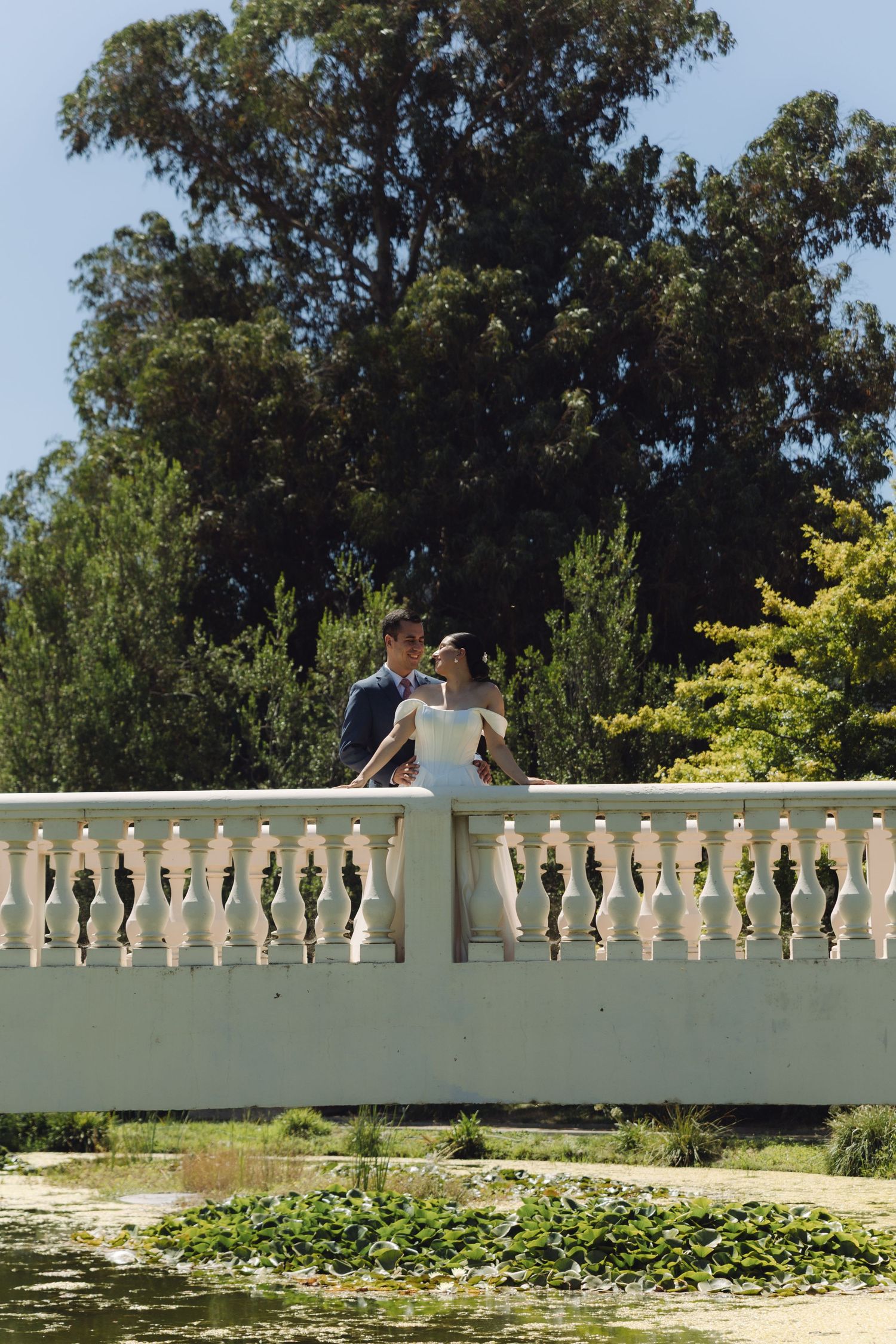 Bride and groom posing on a white balustrade bridge overlooking a serene garden pond.