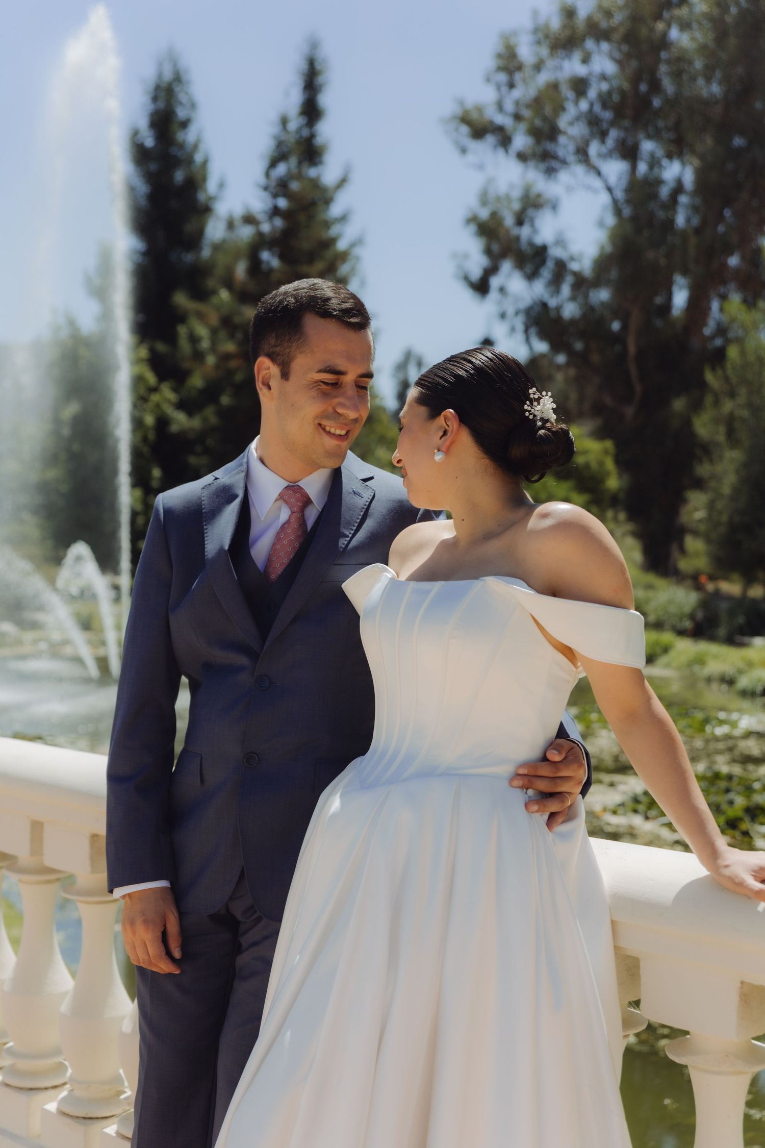 Smiling bride and groom share a tender moment on a balcony with a fountain and lush greenery backdrop.