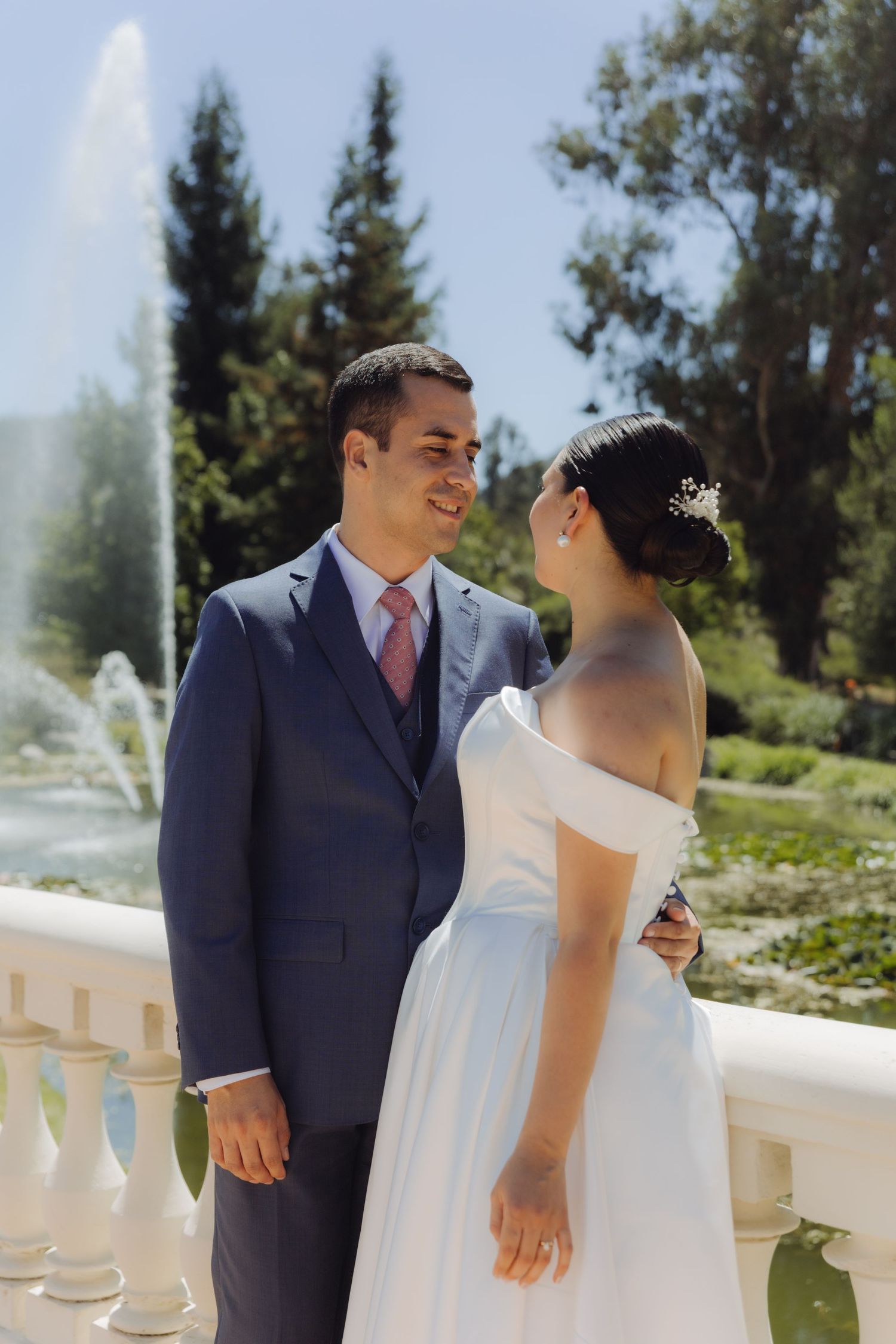 Happy newlyweds gaze at each other lovingly beside a white balustrade with a garden fountain behind them.