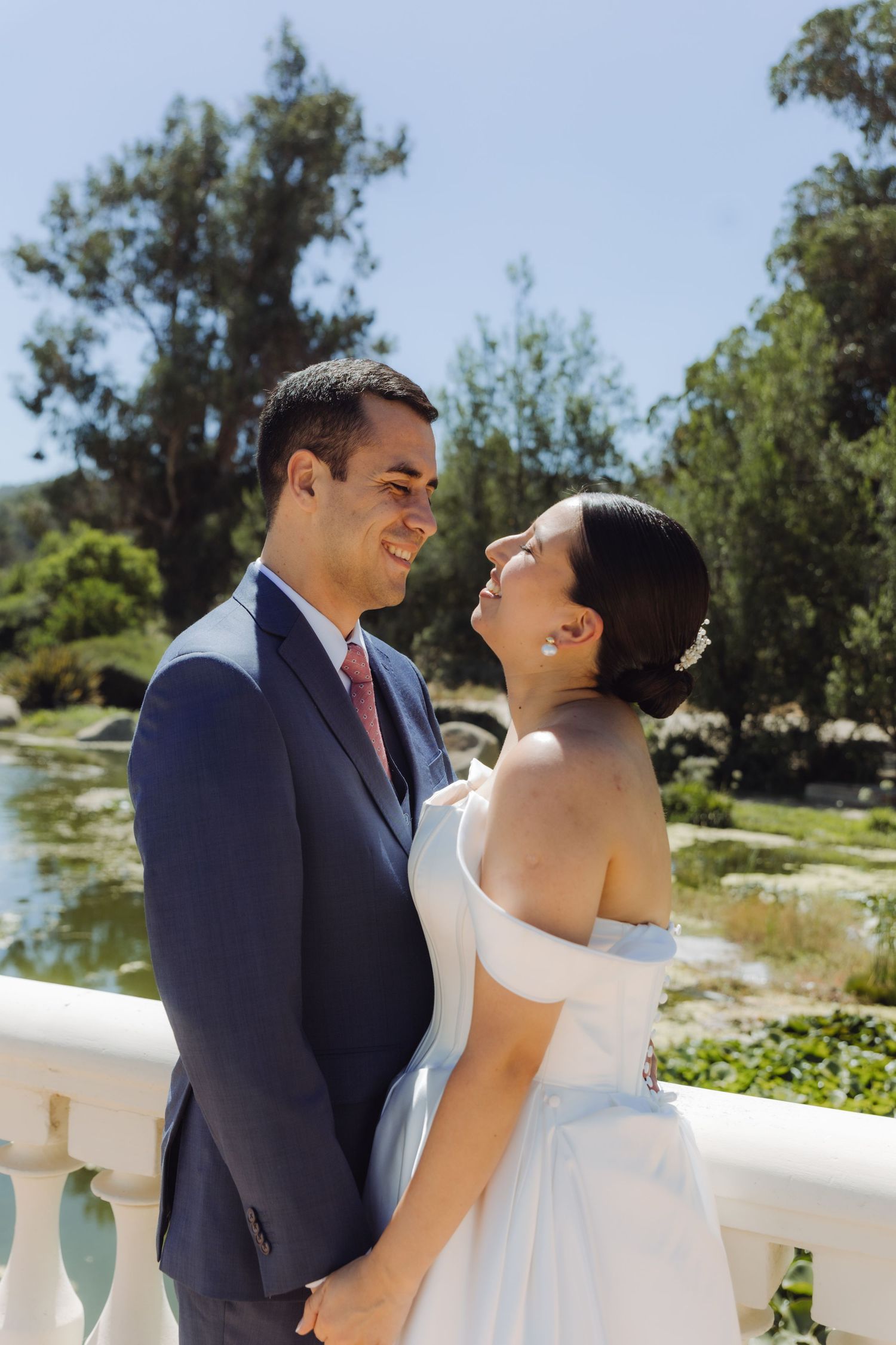 Joyful bride in off-shoulder gown and groom in navy suit embrace near a scenic outdoor fountain.