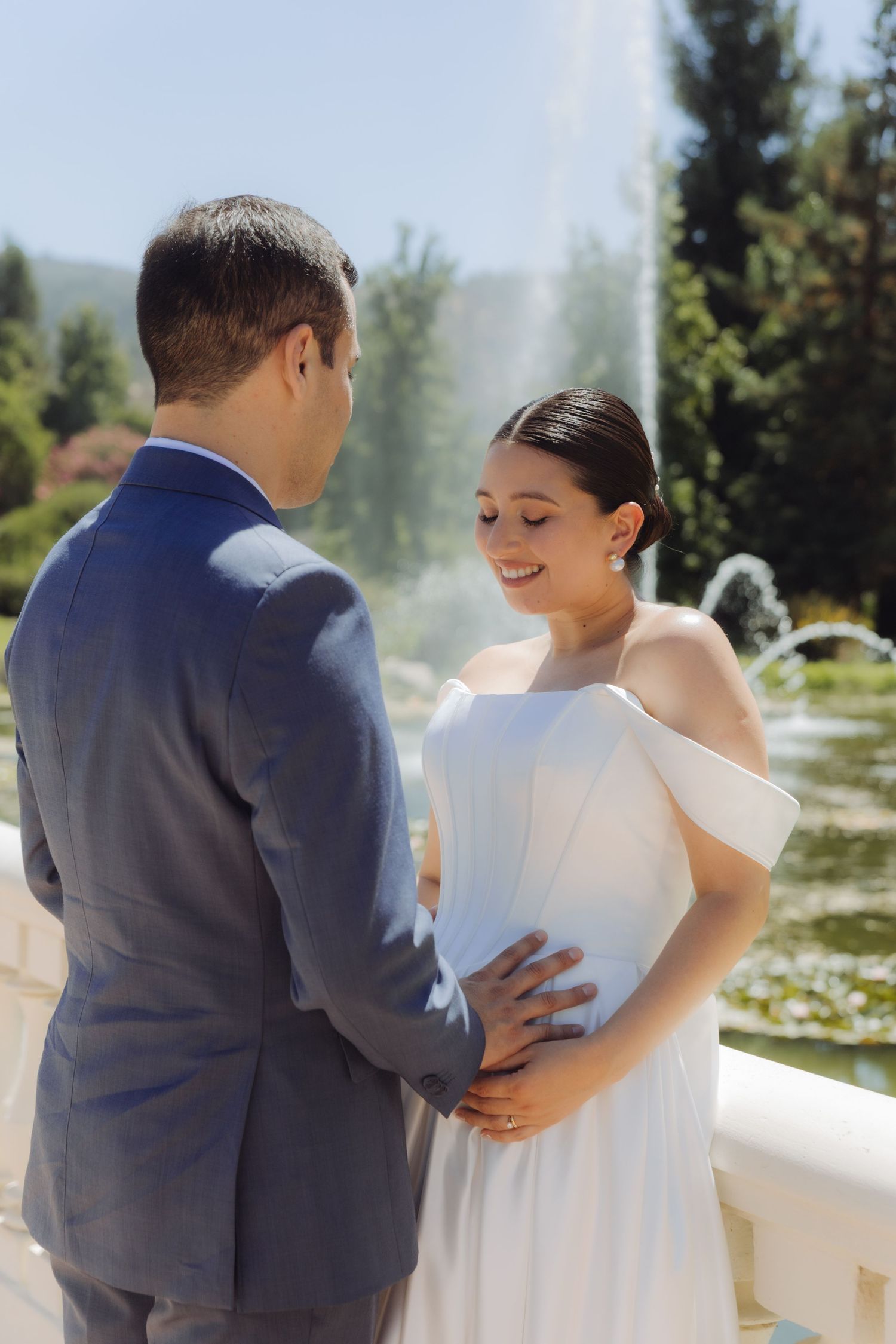 Bride in white off-shoulder dress smiles at groom while holding his waist near a garden fountain.