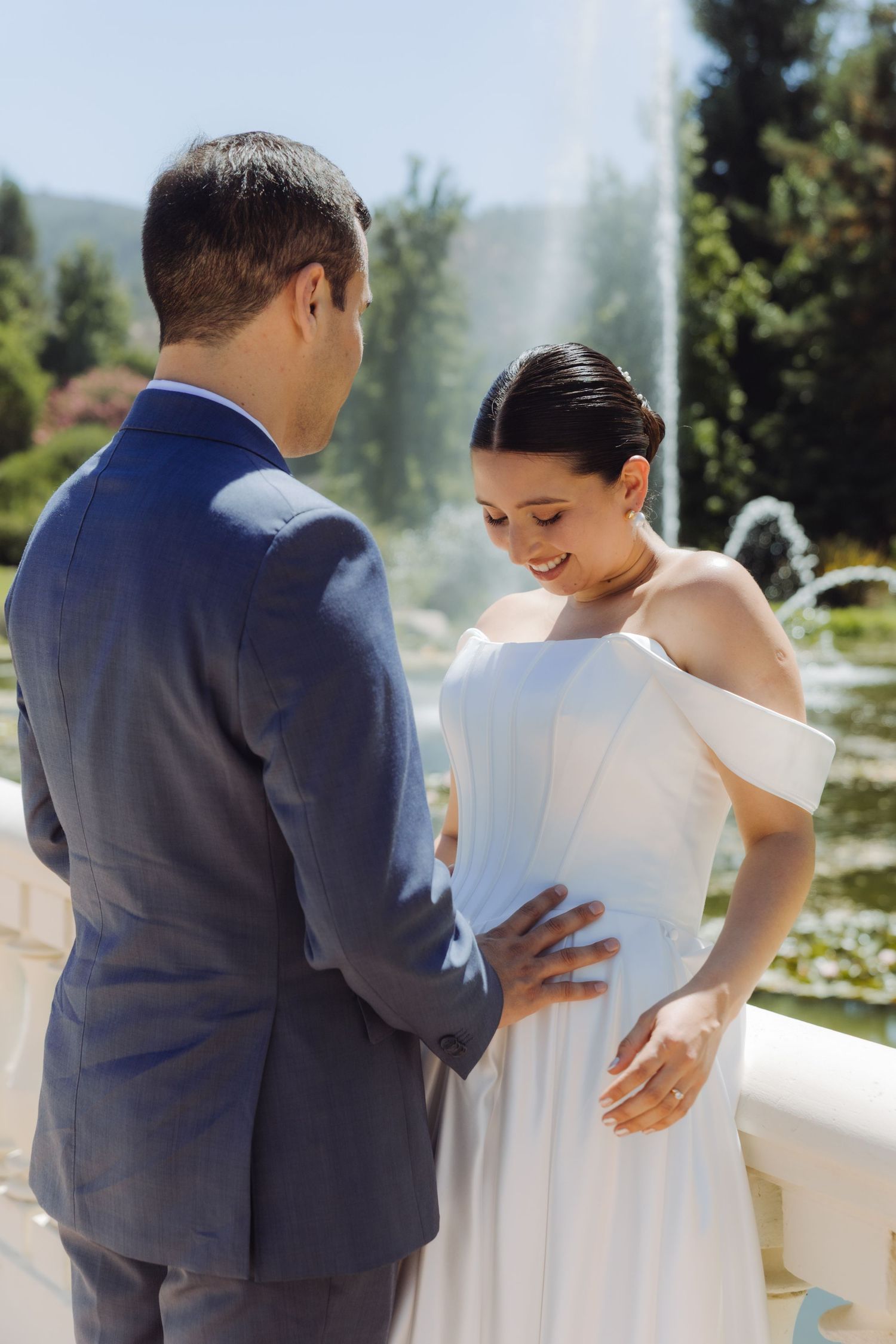 Elegant bride smiles downward as groom places hands on her waist before a lush fountain garden.