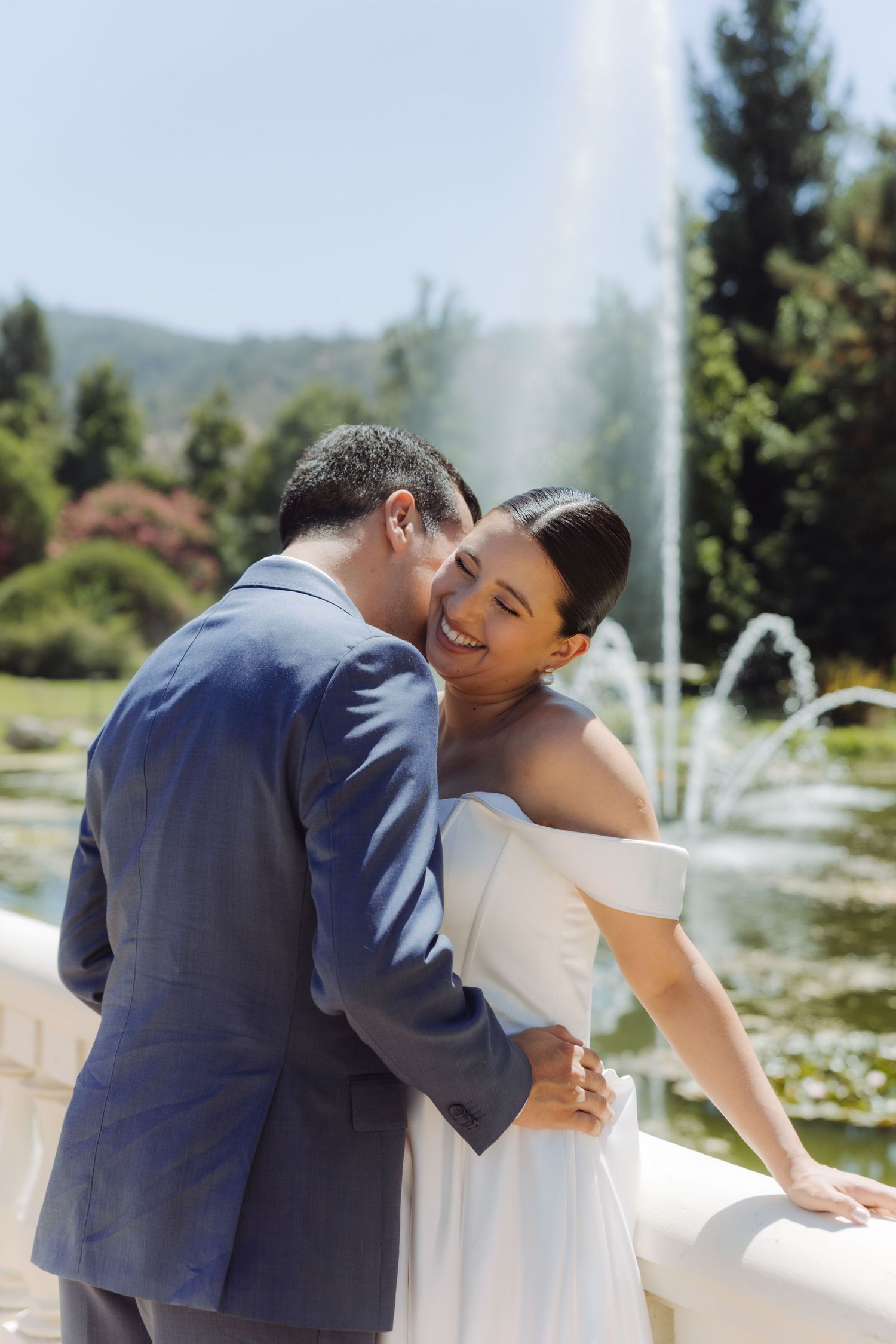 Groom whispers to laughing bride as they embrace by a white railing with a fountain in the background.