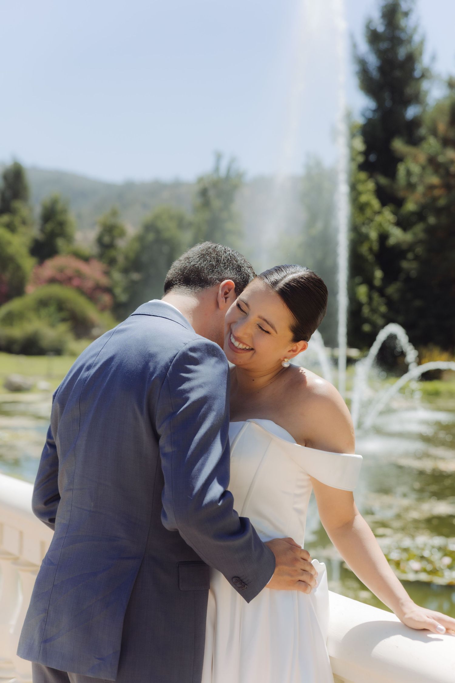 Groom nuzzles smiling bride near a sunlit fountain surrounded by green trees and mountain scenery.