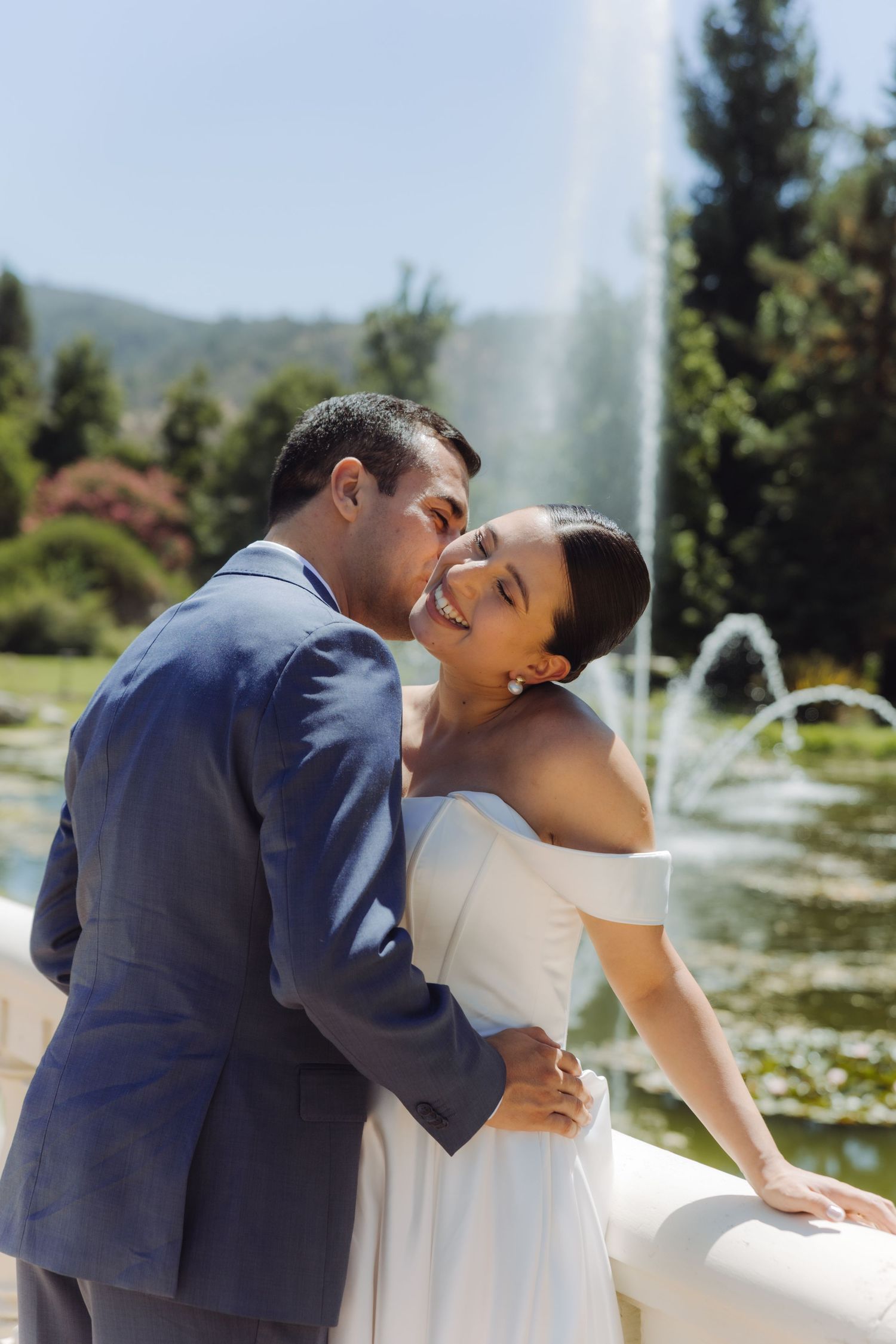 Bride laughs joyfully as groom kisses her cheek beside a sparkling outdoor fountain on their wedding day.
