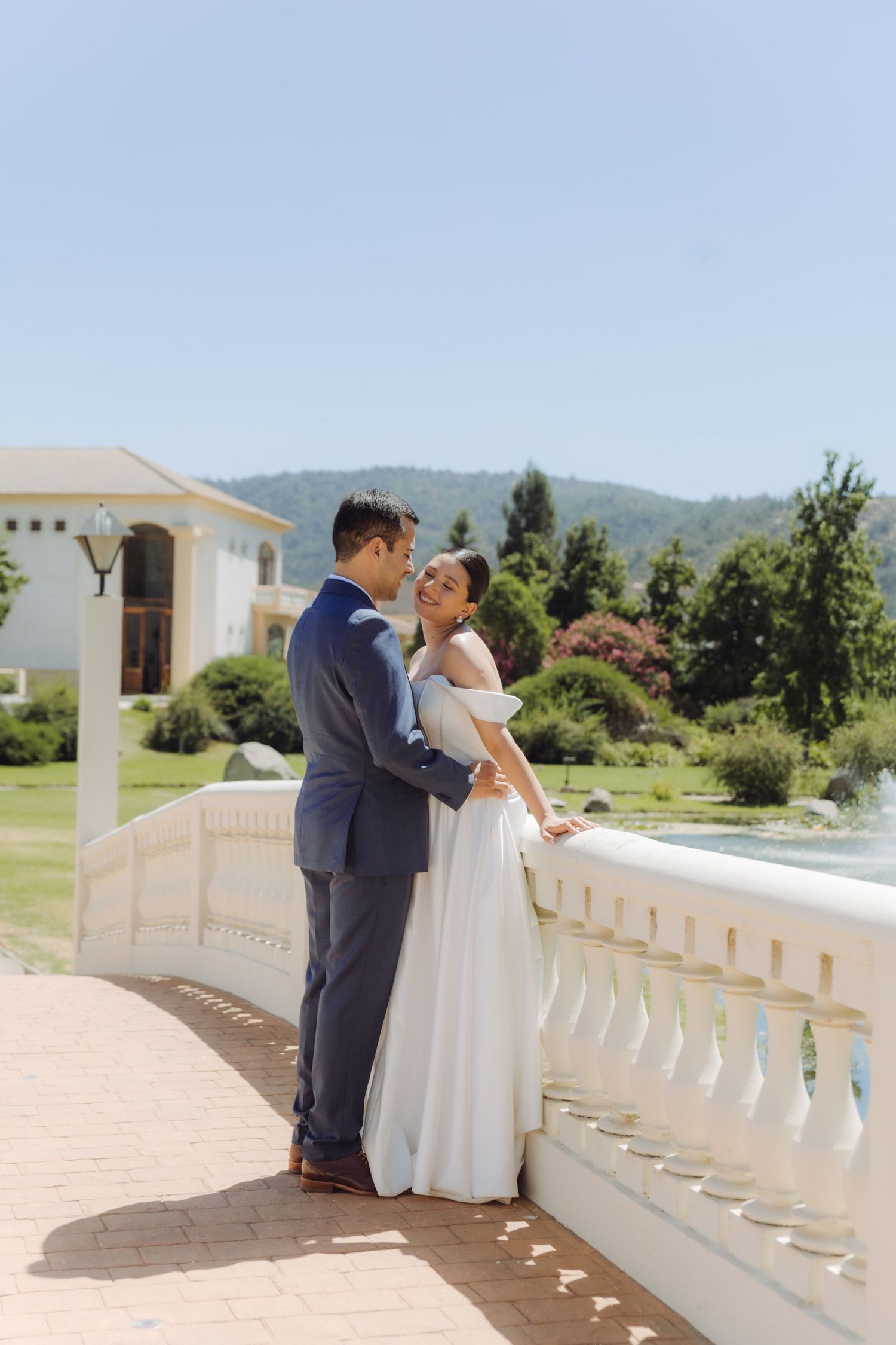 Bride and groom share a romantic embrace on a sunlit terrace with mountains and a fountain behind them.