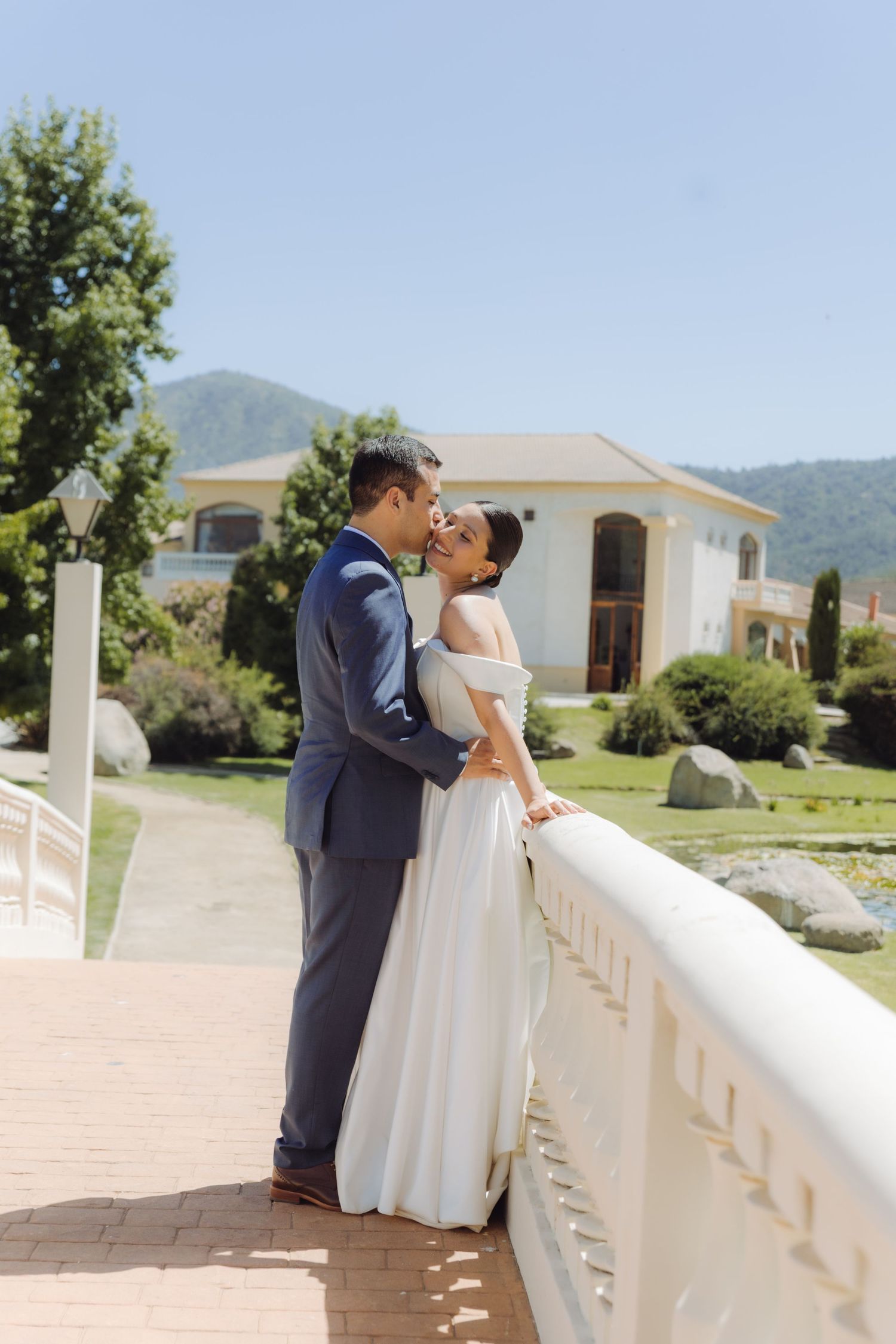 Newlyweds share a kiss on a scenic bridge with a white villa, mountains, and lush garden in the background.