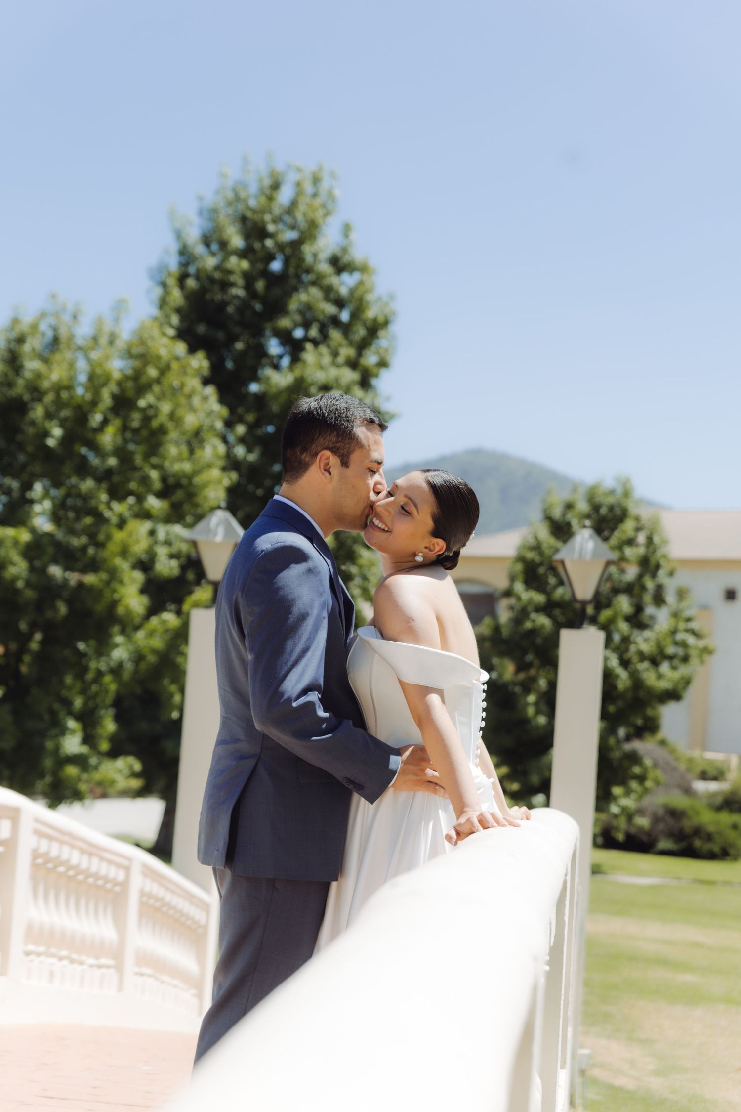 Bride and groom sharing a romantic kiss on a white bridge surrounded by lush greenery on their wedding day.