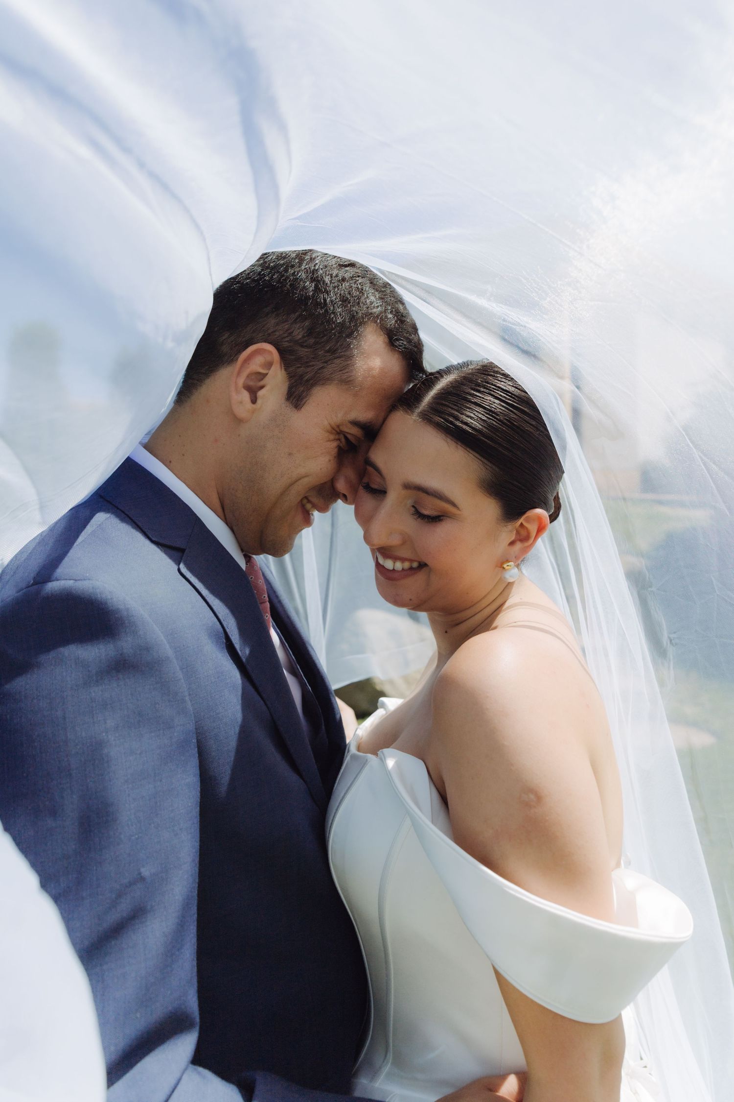 Bride and groom touching foreheads intimately under a flowing white veil on their wedding day.