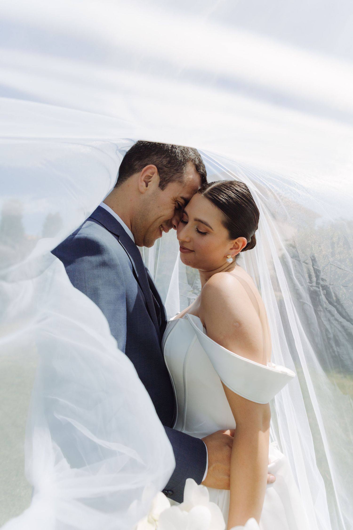Smiling bride and groom embracing under a billowing white veil with a bright sky backdrop.