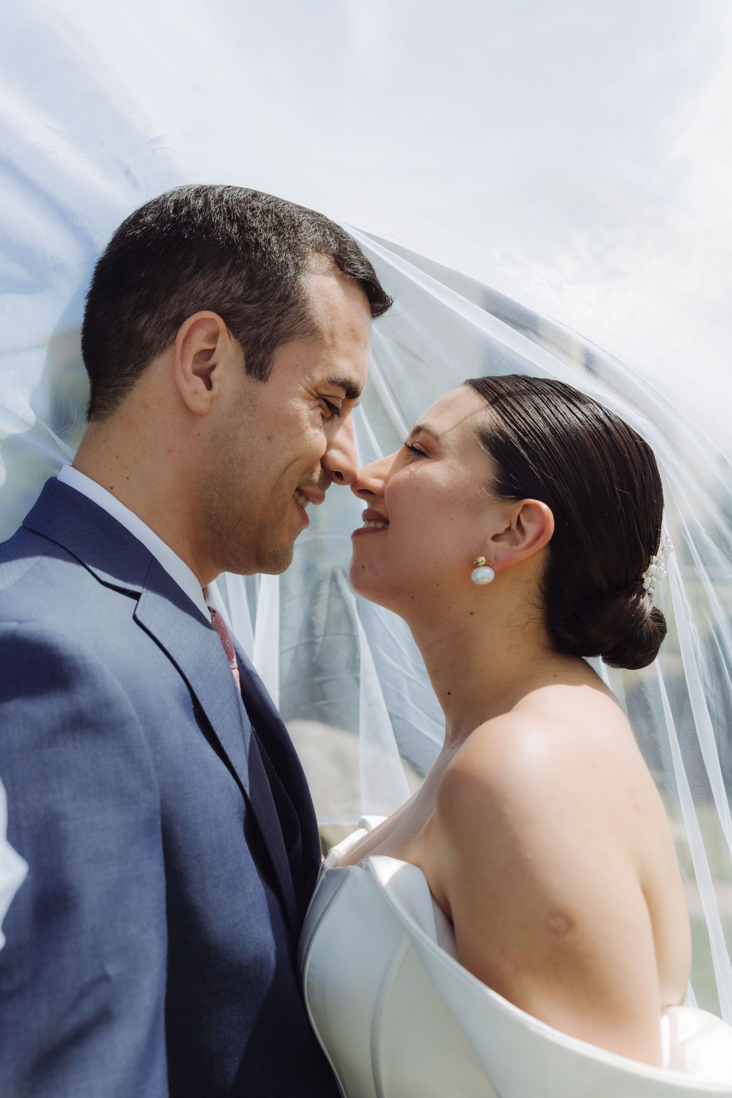 Bride and groom leaning in for a kiss under a white veil, bride wearing pearl earrings.