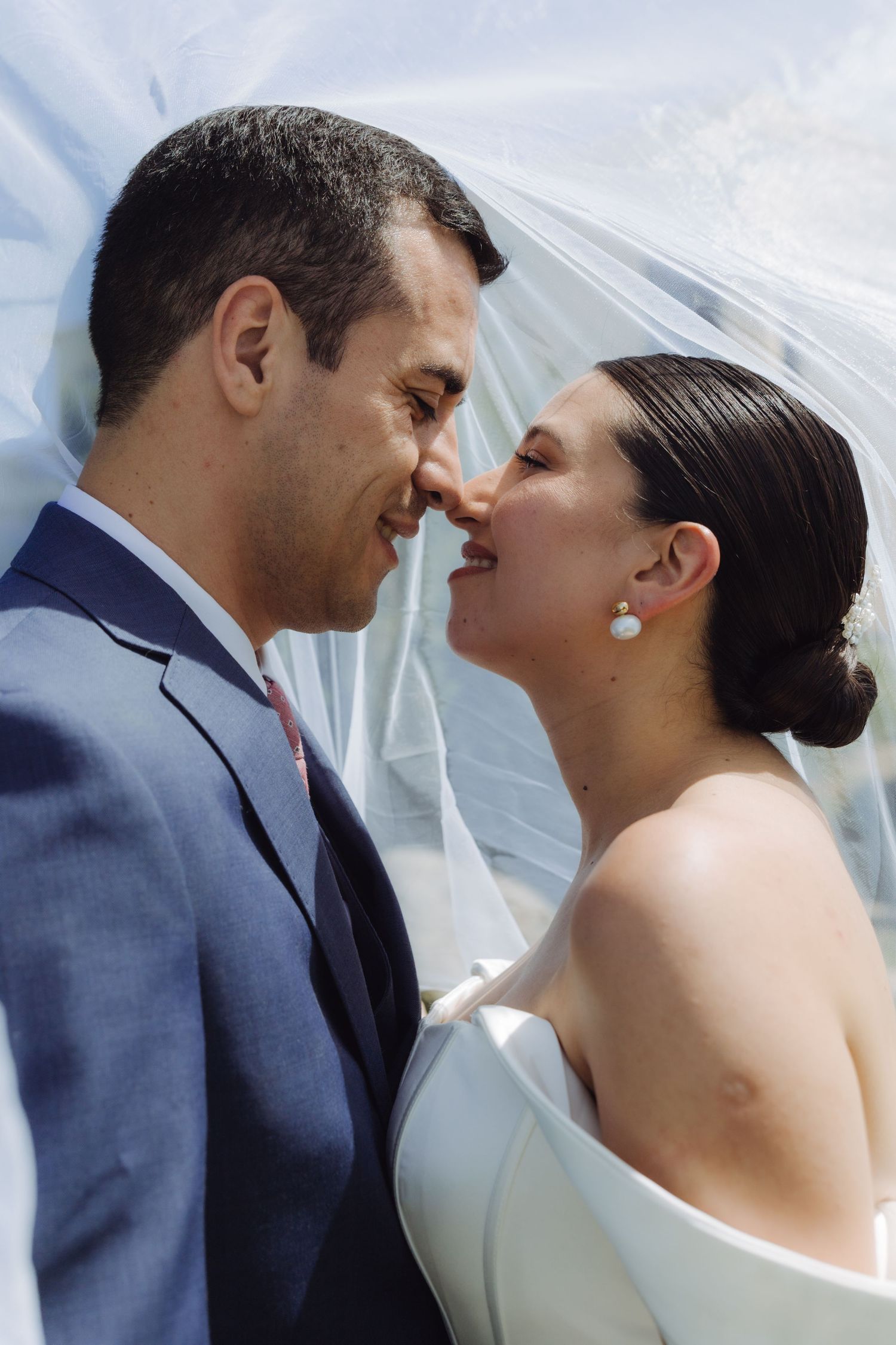 Bride laughing joyfully as she and groom touch noses under a flowing white wedding veil.