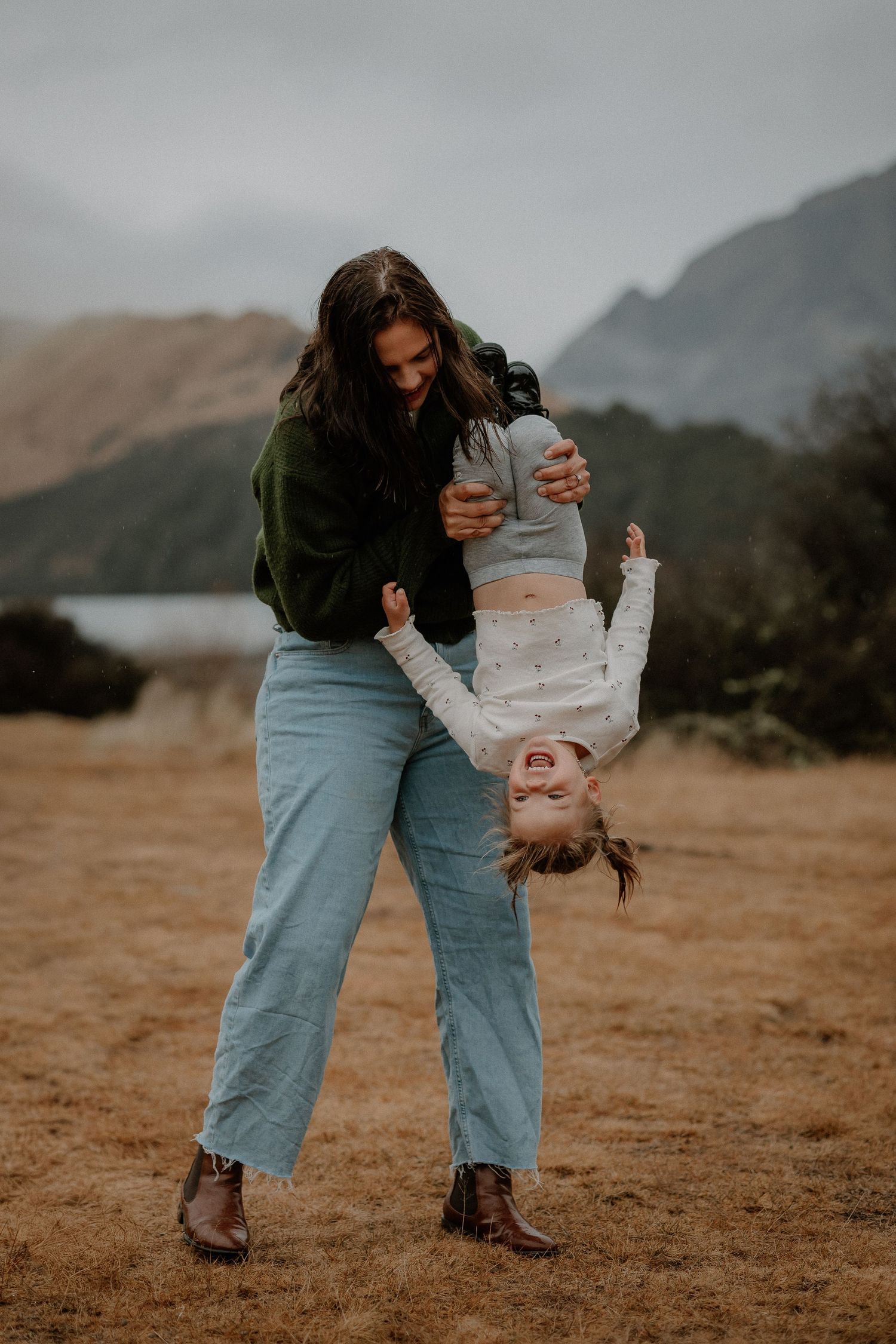 A woman playfully holds a laughing child upside down in a grassy field with mountains in the background.