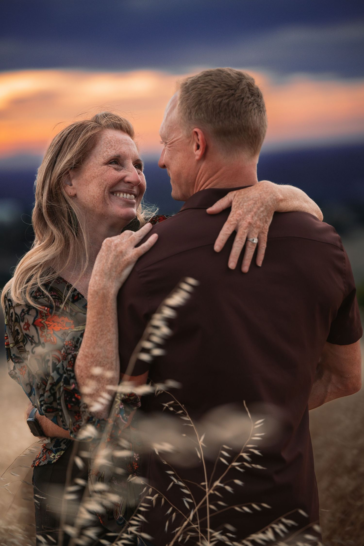 Couple embracing at sunset in a field, woman smiling warmly at man in dark shirt, golden hour sky backdrop.