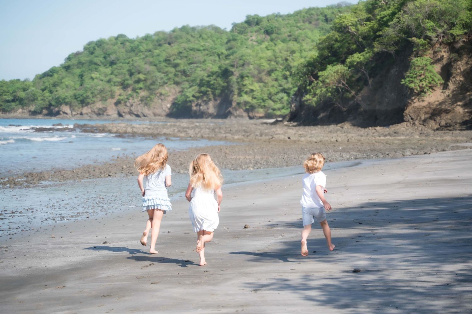 Three girls in white dresses hold hands walking on a tropical beach with lush green hills in the background.