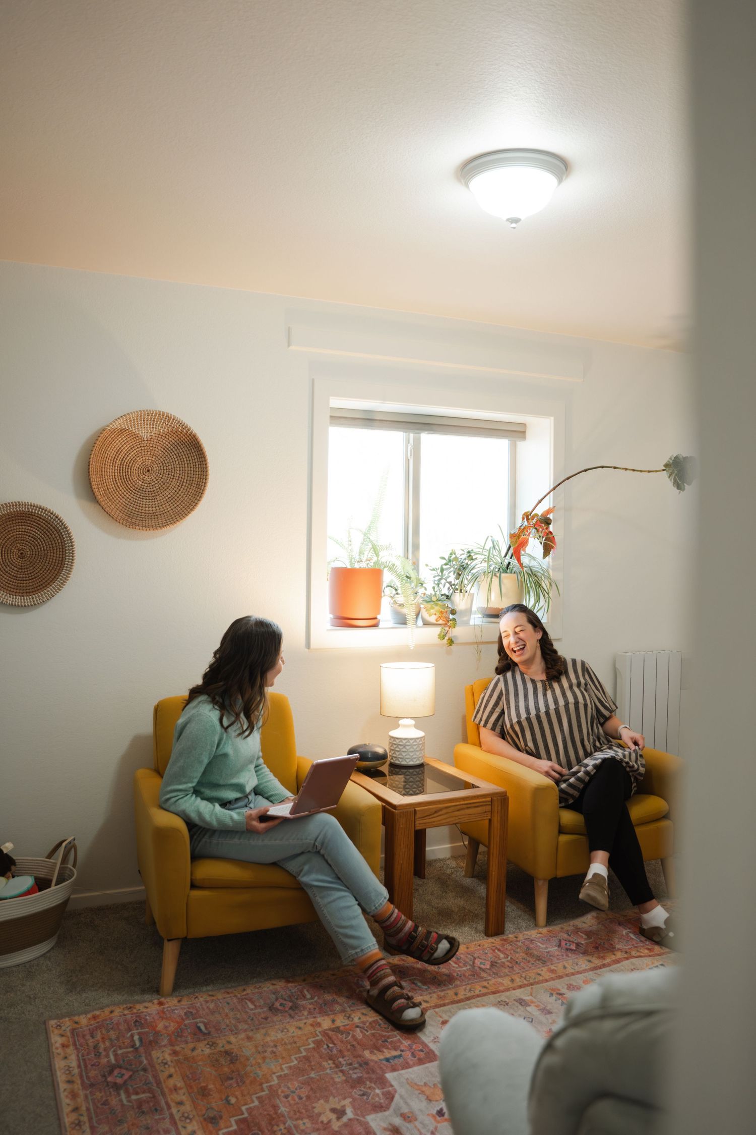 Two women sitting in yellow armchairs having a conversation in a cozy, warmly lit room with plants and woven wall decor.