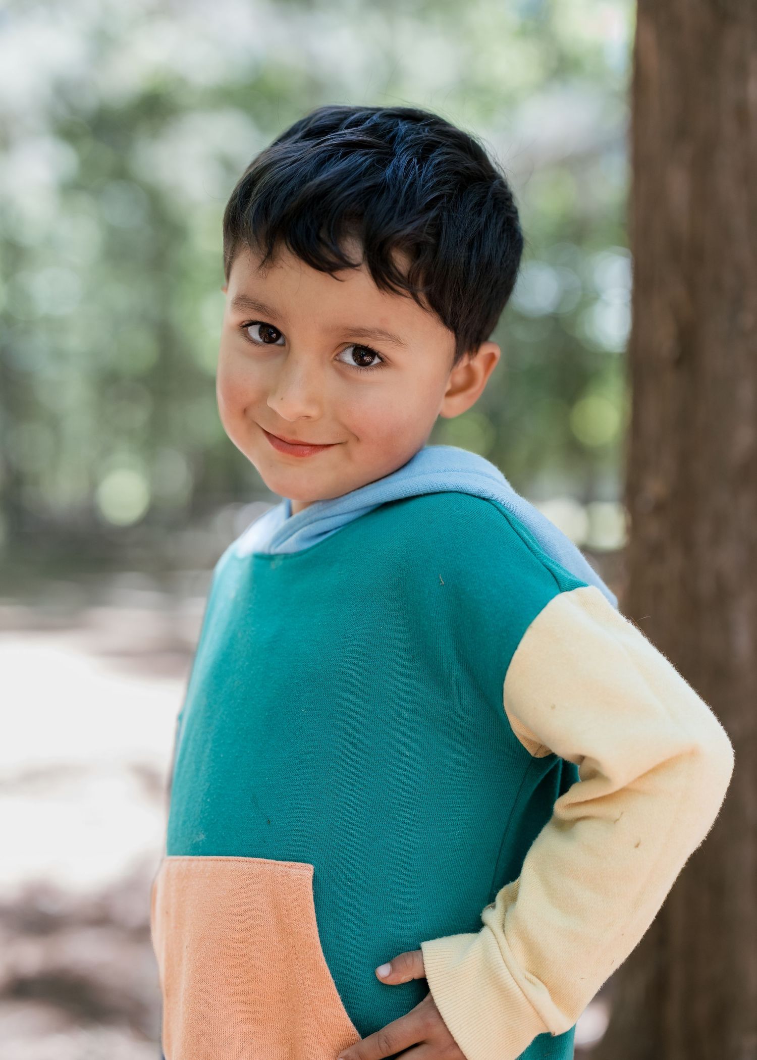 Young boy in colorblock teal, peach, and yellow hoodie smiling over shoulder outdoors near trees.