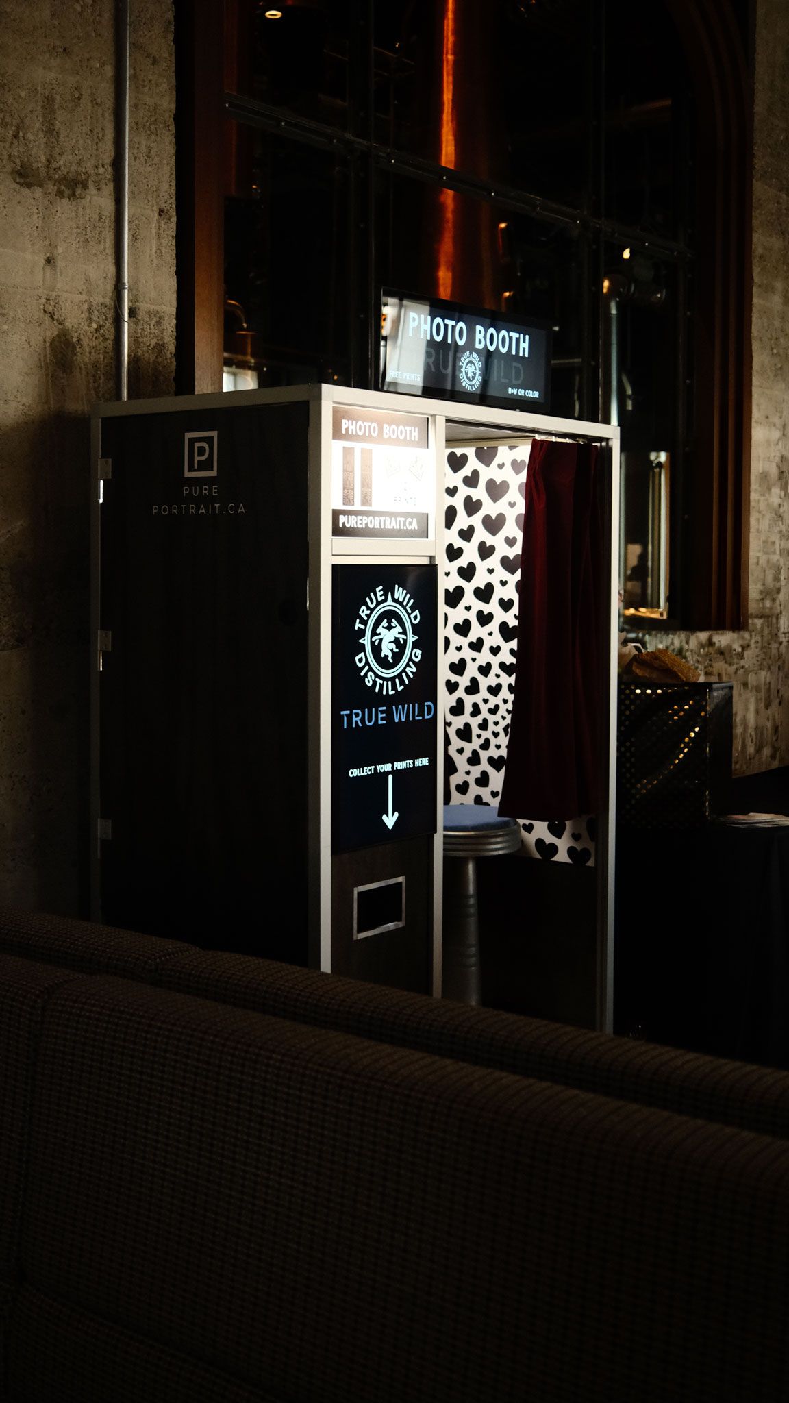 A black photo booth with heart-patterned curtain and True Wild branding displayed in a dimly lit venue.
