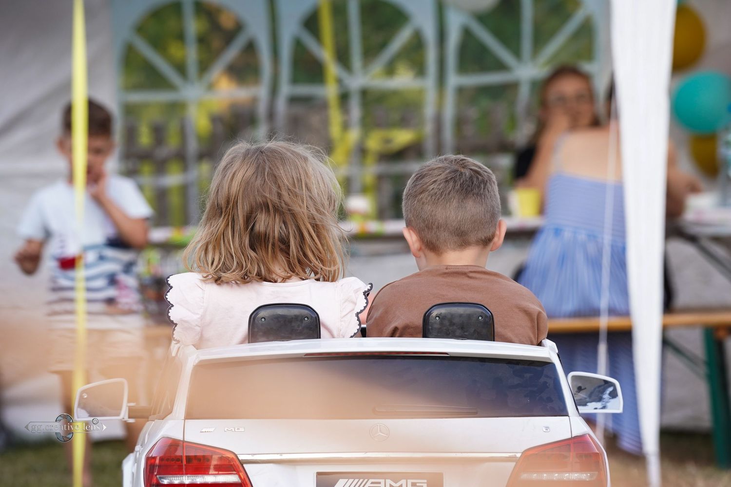 Two children sitting together in a toy electric car at an outdoor garden party.
