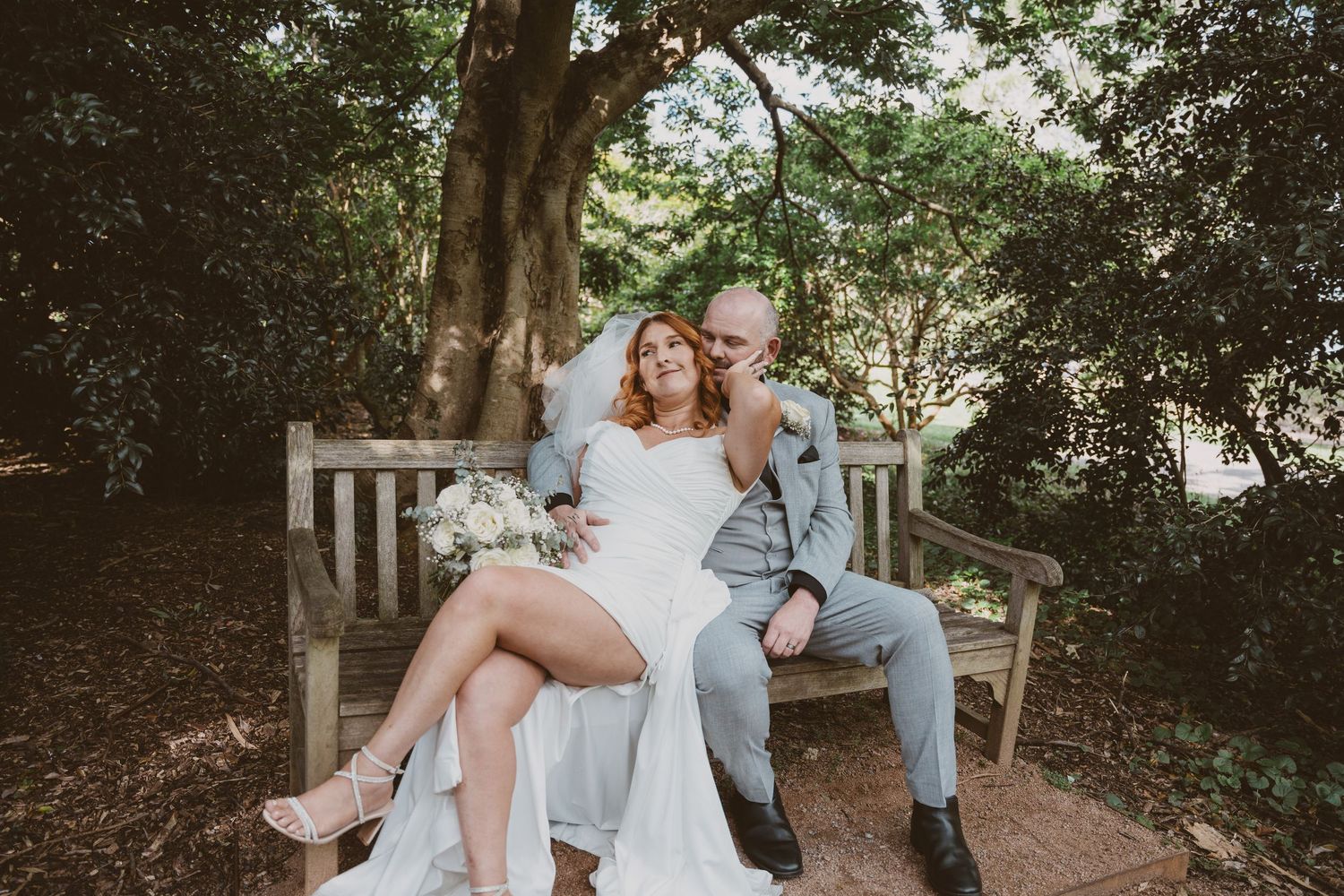 Couple sit together on a wooden bench in a leafy park - relaxed elopement portraits