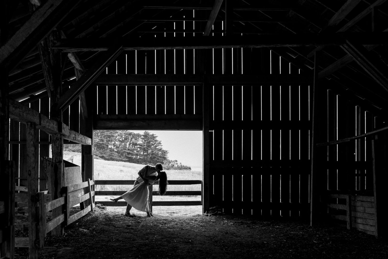 Black and white photo of a couple sharing a dipped kiss inside a rustic wooden barn, silhouetted against the bright open doorway.