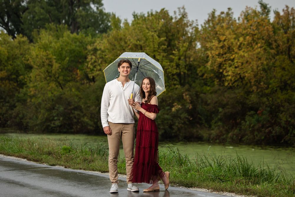 Smiling couple standing close under a clear umbrella on a wet road, her in a long burgundy dress, with green and golden trees behind.