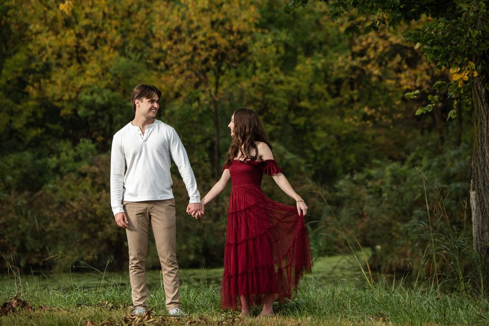A couple holds hands and smiles at each other in a grassy park with autumn trees, she in a burgundy ruffled dress and he in a white sweater.