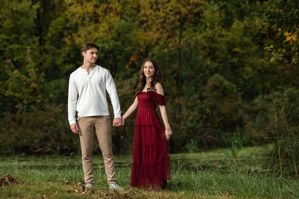 Couple holding hands in a grassy field, he in a white sweater and khakis, she in a flowing red dress, with autumn trees behind.