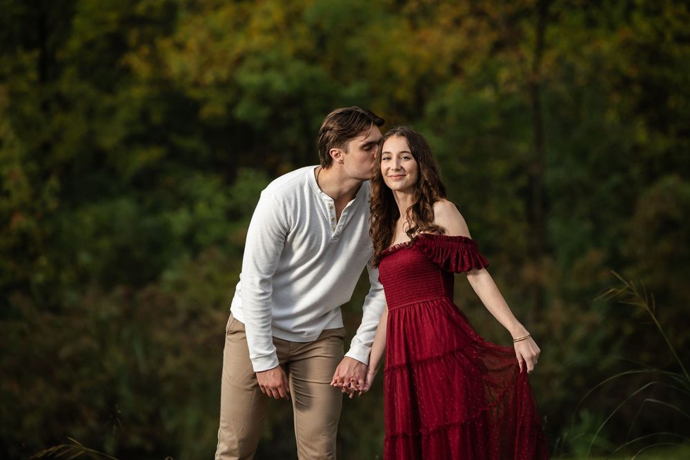 A man in a white sweater and khaki pants kisses the cheek of a smiling woman in a flowing burgundy dress against a blurred autumn forest.