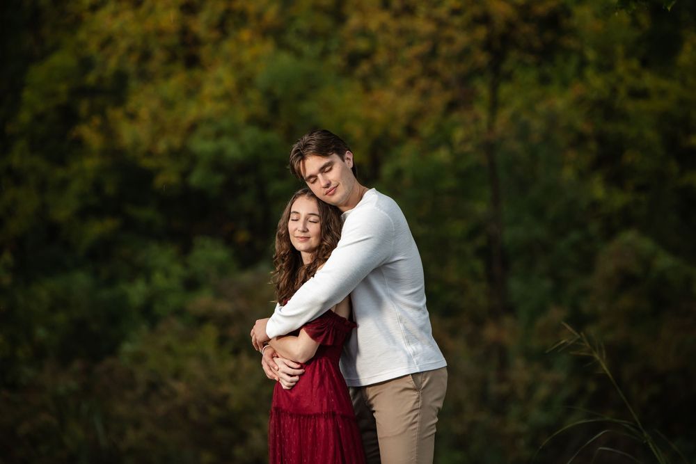 Young couple embracing outdoors, the woman in a red dress and the man in a white sweater, with autumn foliage in the background.