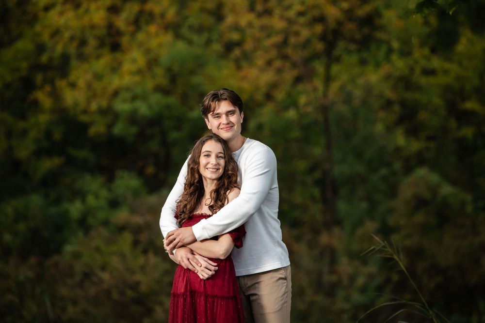 A young man in a white sweater embraces a smiling woman in a red dress from behind against a blurred autumn forest backdrop.