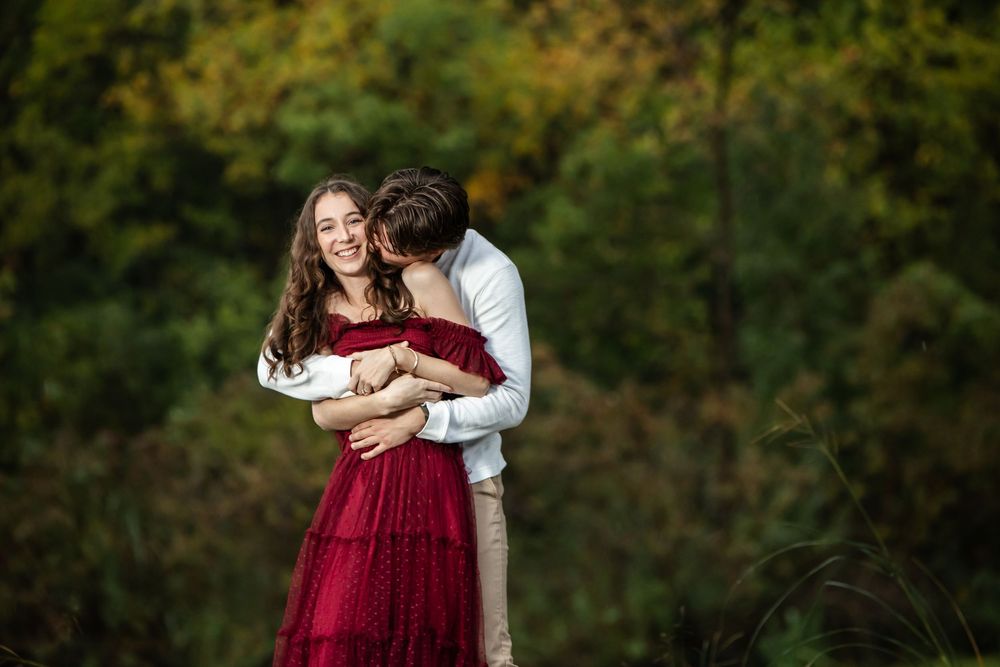 A smiling woman in a flowing red dress is embraced from behind by a man kissing her cheek in front of colorful autumn trees.