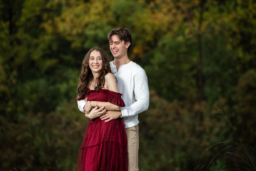 Smiling couple embracing outdoors, a woman in a flowing burgundy off-the-shoulder dress held by a man in a white shirt against autumn trees.