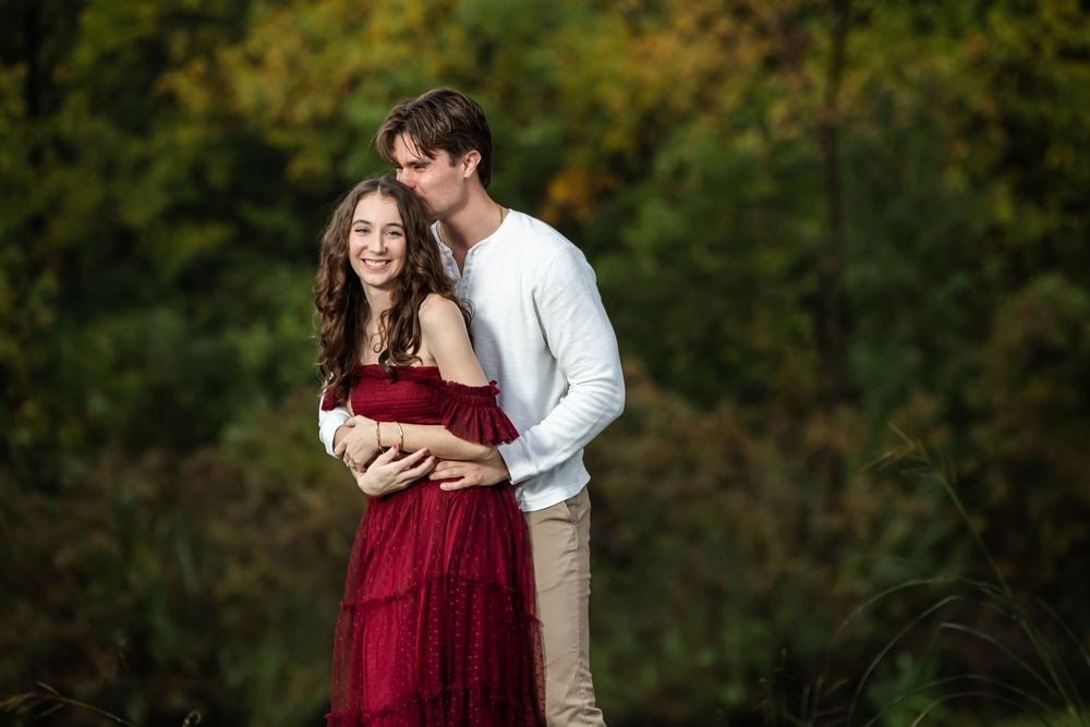 A smiling woman in a burgundy off-the-shoulder dress embraces a man in a white sweater and khaki pants against an autumn forest backdrop.