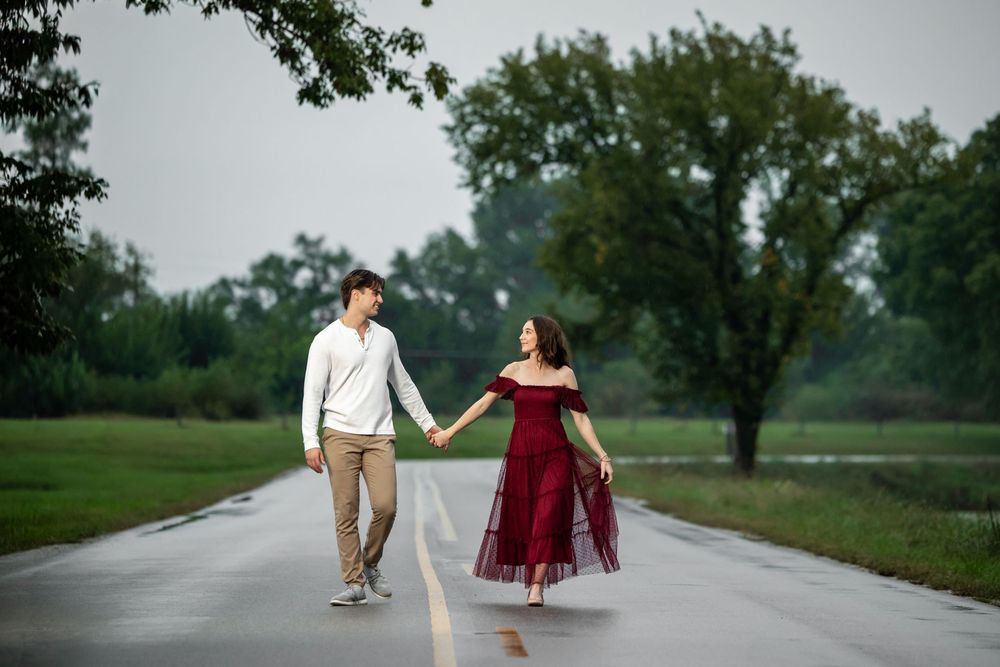 Couple holding hands walking down a wet country road, she wears a flowing red dress and he a white shirt, with green trees beyond.