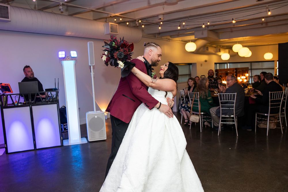 Bride in a white gown and groom in a burgundy suit share a first dance at a wedding reception while guests watch.