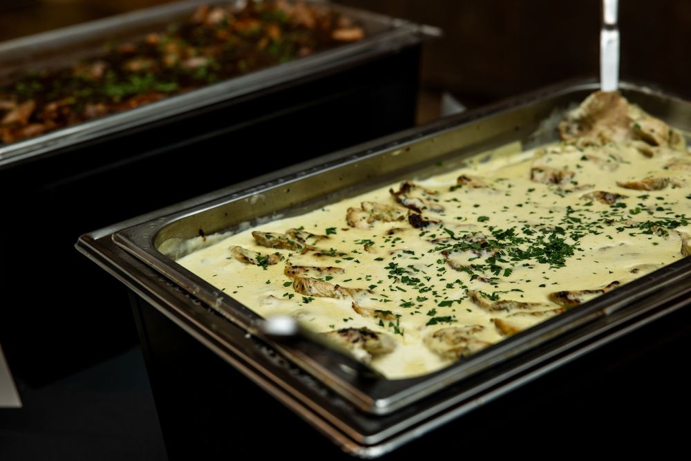 Chafing dish filled with creamy mushroom sauce topped with fresh chopped herbs on a buffet table, with another tray of food behind it.