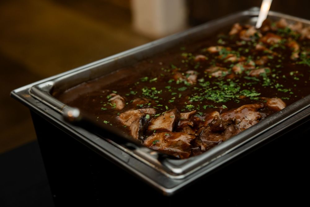 Stainless steel chafing dish filled with tender braised meat in rich brown gravy, garnished with fresh chopped parsley at a buffet.