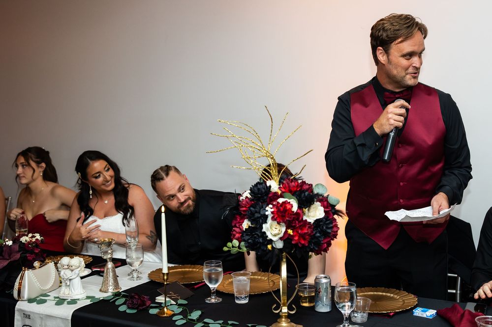 Man in burgundy vest gives a wedding toast with a microphone beside a red and white floral centerpiece as guests smile at the head table.