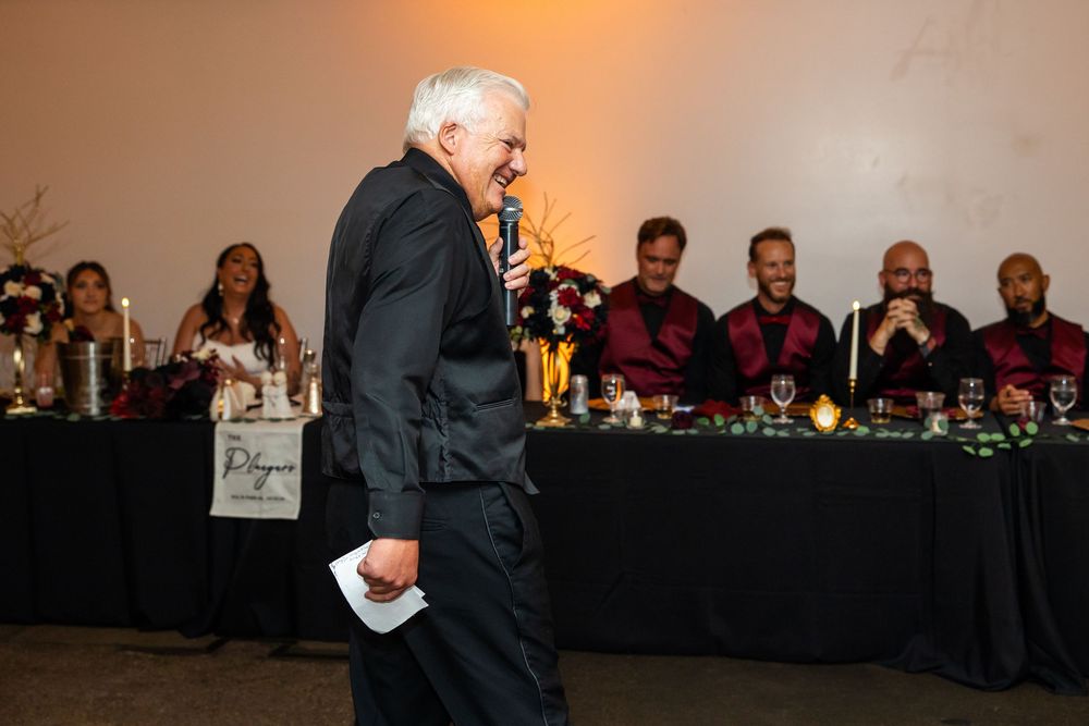 Gray-haired man in black holding a microphone and paper gives a speech at a reception while seated guests in burgundy shirts smile.