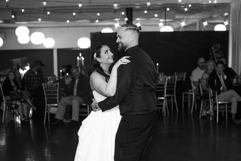 Bride in a white gown and groom in a black shirt share a slow dance on the floor at a wedding reception while guests watch.