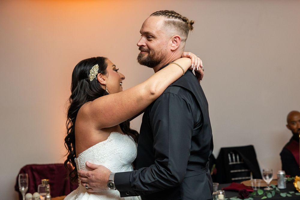 A smiling bride in a white strapless gown and a bearded groom in a black shirt share a close wedding dance embrace.