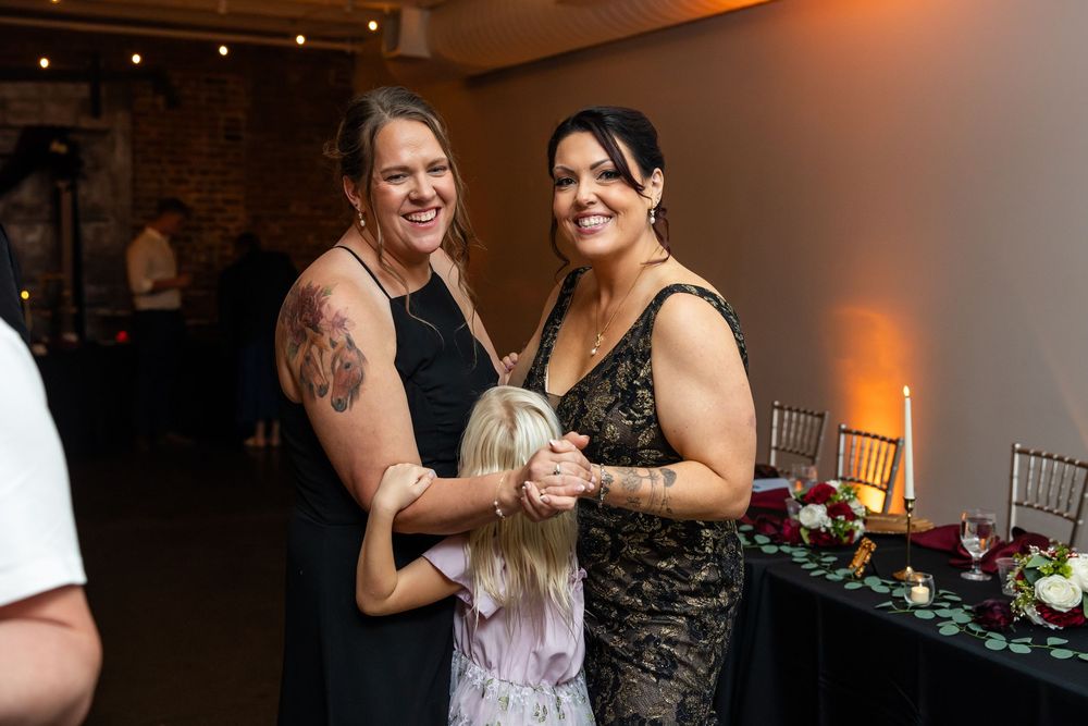 Two smiling women in black dresses hold hands with a young girl at an elegant reception with candlelit tables and floral centerpieces.