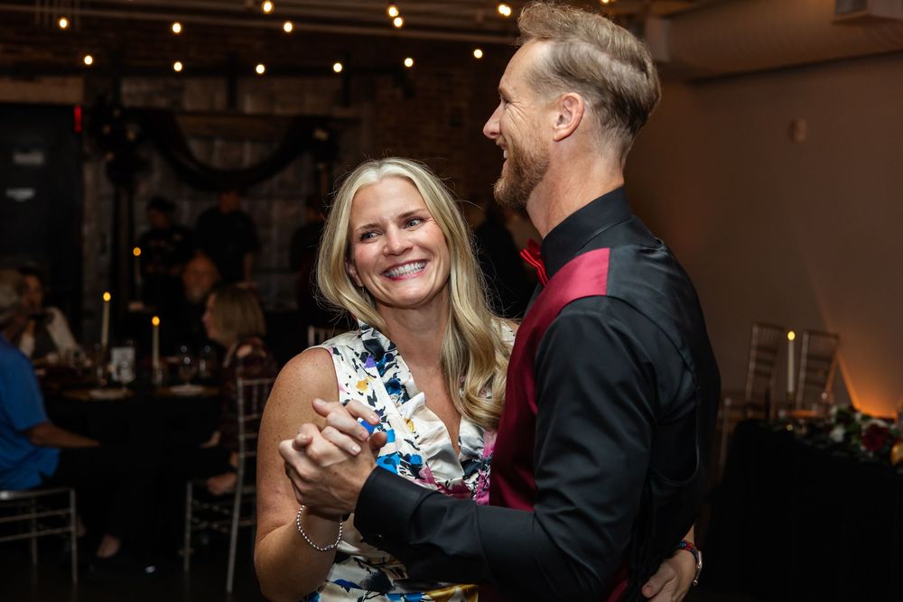 A smiling blonde woman in a floral dress dances with a man in a black shirt at an evening reception with string lights.