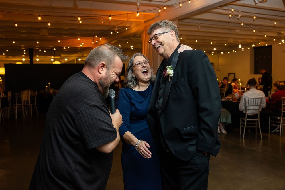 Three people laugh together at a wedding reception as one man speaks into a microphone beneath warm string lights.