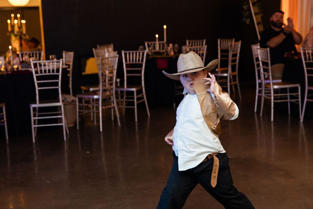 Performer in a cowboy hat and white shirt dances on a polished floor at an event venue with candlelit tables and chairs behind.