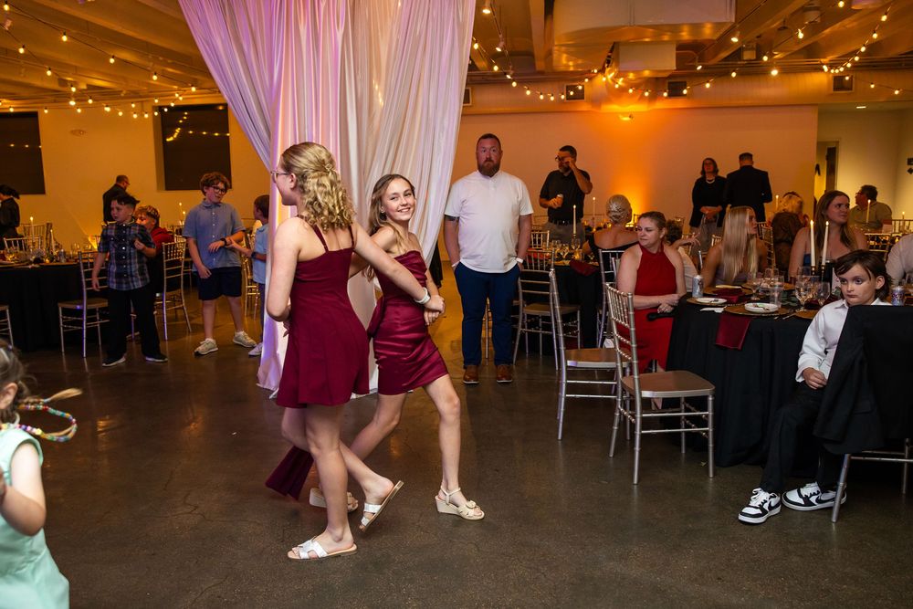 Two women in burgundy dresses walk across the dance floor at an indoor wedding reception with string lights and guests seated at tables.