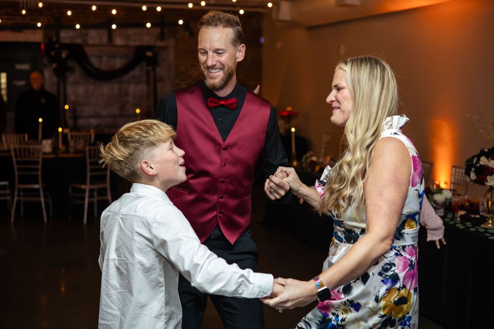 Smiling man in a burgundy vest and bow tie dances with a young boy and a blonde woman in a floral dress at an indoor wedding reception.