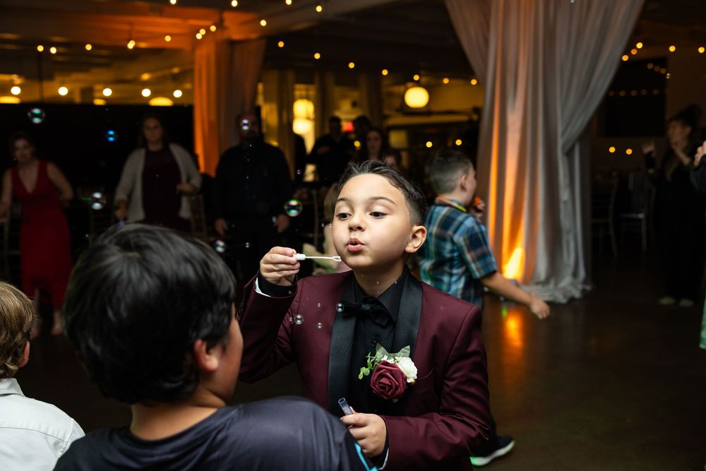 Boy in a burgundy suit with a floral boutonniere blows bubbles at a lively indoor wedding reception while guests mingle in the background.